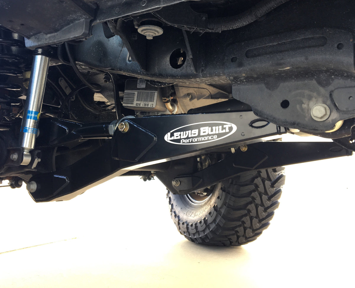 Close-up of a leveled Ford Super Duty undercarriage showing a Lewis Built Performance adjustable radius arm with a visible brand sticker, suspension components, and part of a tire against a white background.