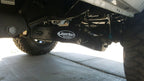 Close-up of a leveled white Ford Super Duty undercarriage featuring Lewis Built Performance logo badges on adjustable radius arms, photographed on a concrete surface.