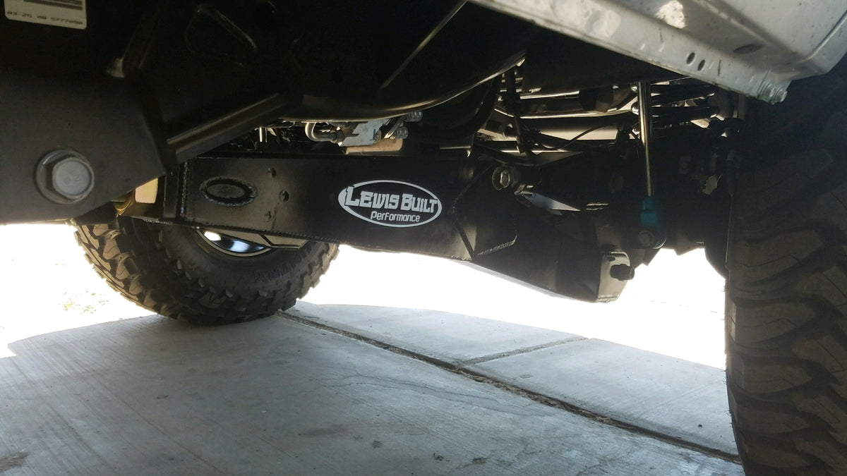 Close-up of a leveled white Ford Super Duty undercarriage featuring Lewis Built Performance logo badges on adjustable radius arms, photographed on a concrete surface.