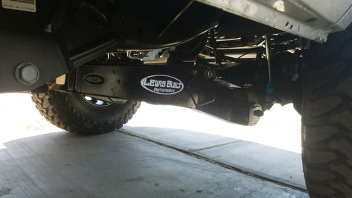 Close-up of a leveled white Ford Super Duty undercarriage featuring Lewis Built Performance logo badges on adjustable radius arms, photographed on a concrete surface.