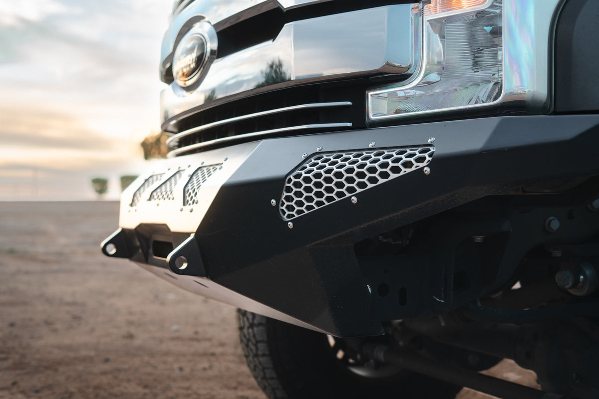 Close-up of a black Lewis Built Performance STRYKER front bumper with a honeycomb grille insert and recovery points on a leveled Ford Super Duty in a desert setting at sunset.