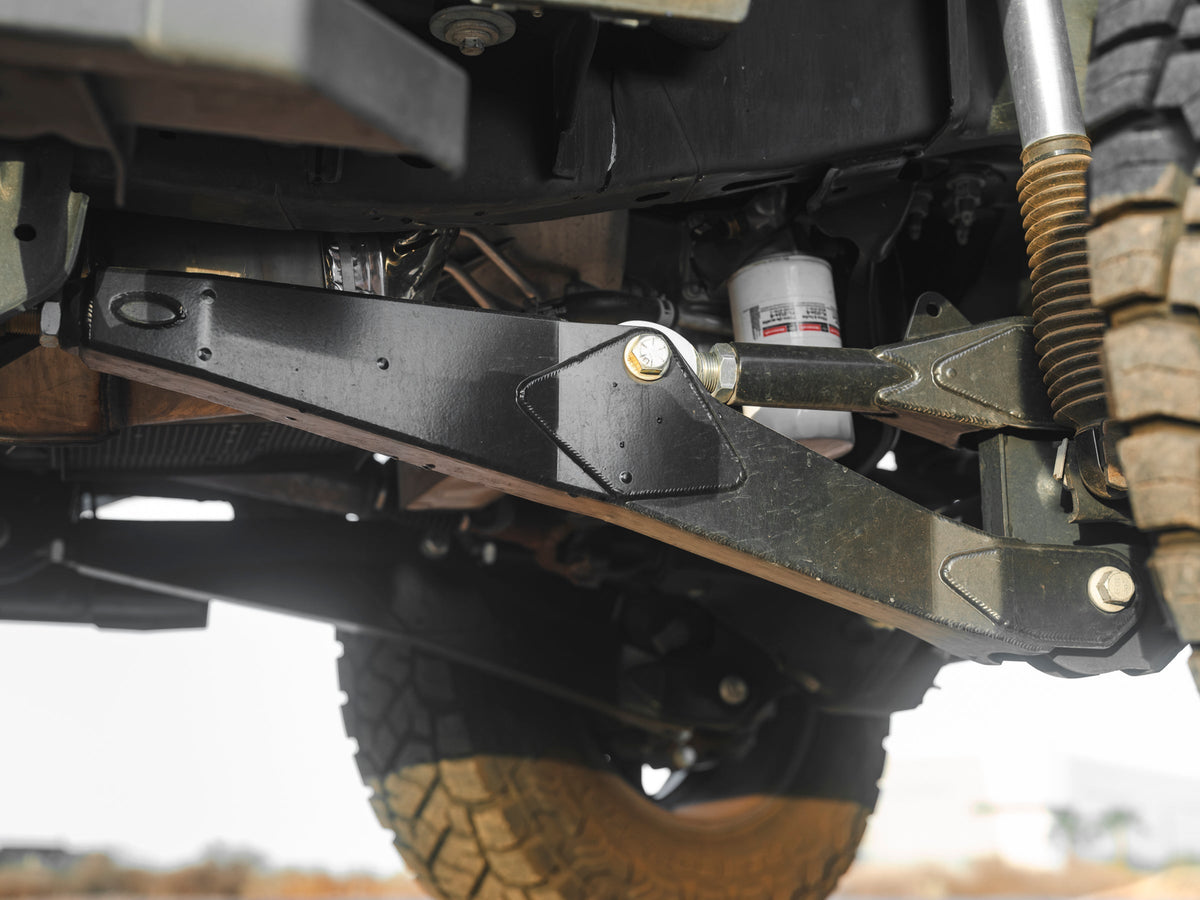 Close-up of heavy-duty Lewis Built Performance Ford Super Duty radius arms connected with Grade 8 bolts and rod ends, with aftermarket shock absorbers visible, set against reddish-brown dirt.