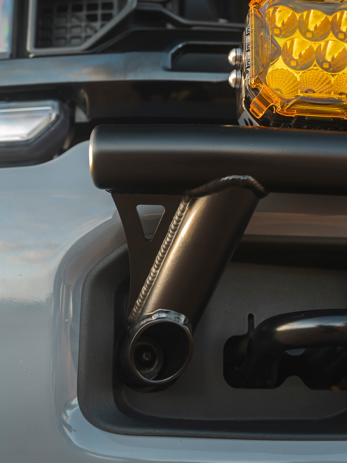 Close-up of a black Lewis Built Performance APEX bumper light mount installed on a leveled gray Ford Super Duty, with part of a bright amber off-road light visible above.