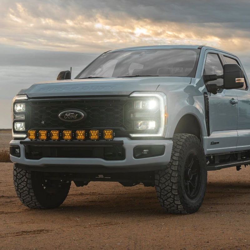 Leveled gray Ford Super Duty pickup truck featuring a black grille, auxiliary LED lights, and a Lewis Built Performance APEX bumper light mount, parked on a dirt road under a dramatic sky.