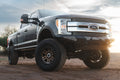 Dark gray lifted Ford Super Duty truck with large off-road tires and a black Lewis Built Performance STRYKER front bumper, photographed on dirt ground beneath a cloudy sky with warm sunlight.