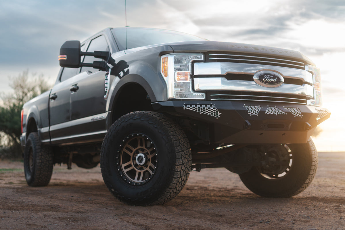 Dark gray lifted Ford Super Duty truck with large off-road tires and a black Lewis Built Performance STRYKER front bumper, photographed on dirt ground beneath a cloudy sky with warm sunlight.