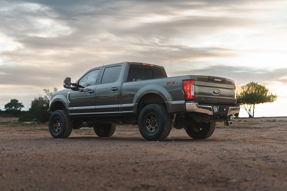 A lifted gray Ford Super Duty pickup truck with off-road tires, parked on a dirt road under a cloudy sunset sky.