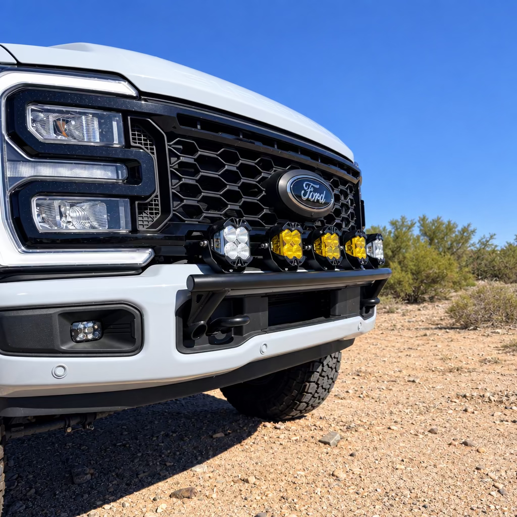A close-up outdoor shot of the front bumper of a gray Ford Super Duty on a dirt road under a clear blue sky. The truck is equipped with a Lewis Built light mount with amber Baja Designs LP4 lights.