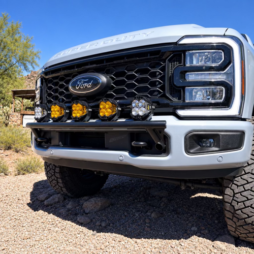 A close-up front view of a light blue Ford truck parked outdoors on a dirt surface under a clear blue sky. The truck has a black front grille, black bumper, and four yellow auxiliary lights mounted across the grille.